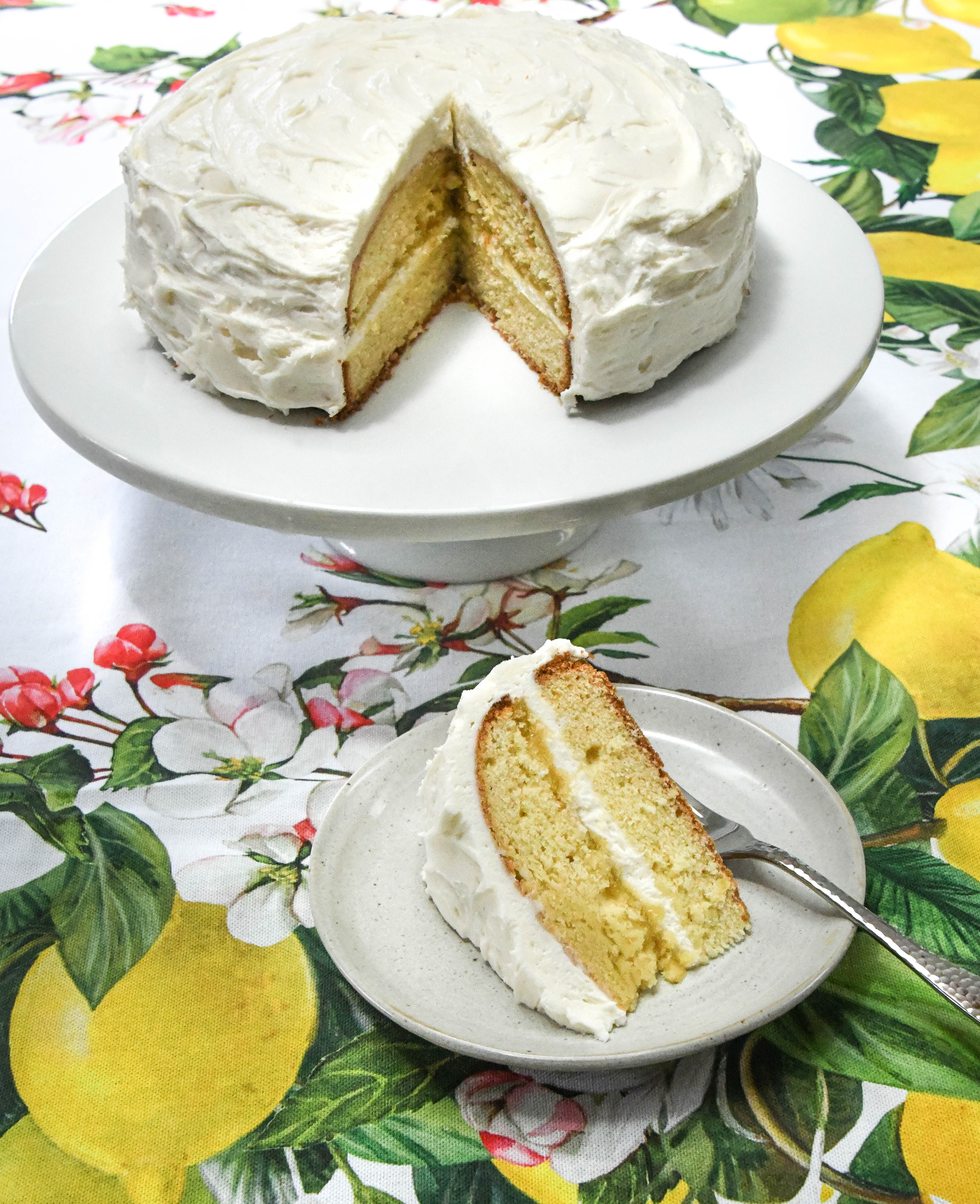 Sliced lemon curd cake on plate and cake stand. Both are on a lemon-decorated tablecloth